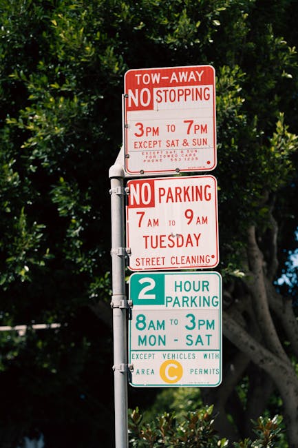 Close-up of a metal parking restriction signpost positioned against a stone wall in an urban environment. The top sign indicates no parking except by permit from 7am to 6pm, Monday to Friday, with a r