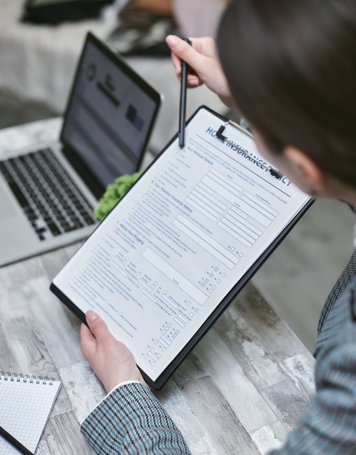 A person wearing glasses and a checkered blazer is seated at a wooden table, holding a clipboard with a filled-in liability and insurance form for house removals. The individual is using a pen to revi