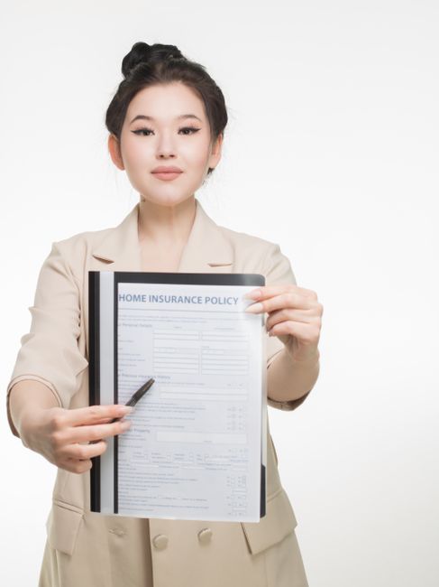 A person wearing glasses and a checkered blazer is seated at a wooden table, holding a clipboard with a filled-in liability and insurance form for house removals. The individual is using a pen to revi